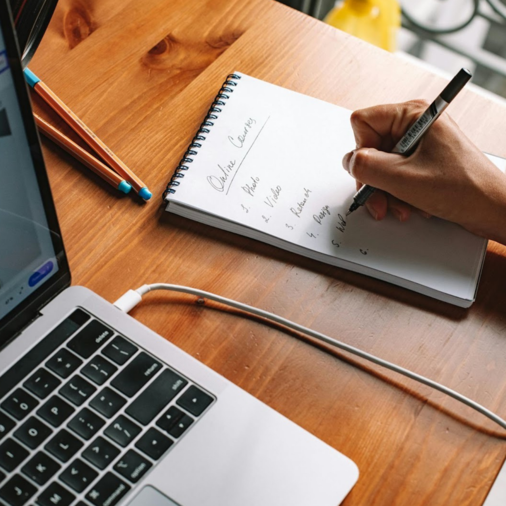 Person Writing on a Notepad Beside a Laptop